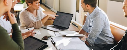 Manager pointing at laptop screen and talking to colleagues during business meeting. Female executive showing data to team in meeting.